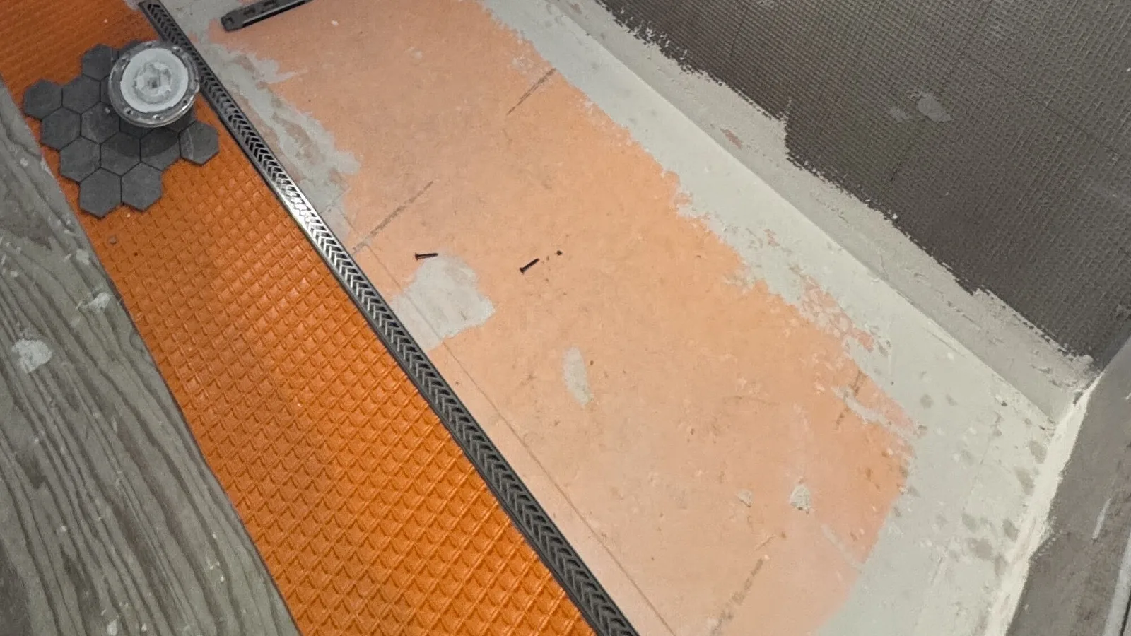 Shower floor prepped with an orange waterproofing membrane and a stainless linear drain, with mosaic tile stacked ready to set against cement board walls