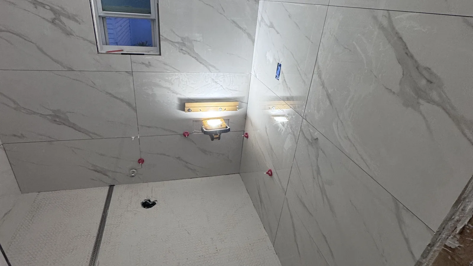 Shower walls tiled in large-format marble-look porcelain around a window, lit by work lights, with a white mosaic floor in a St. Petersburg bathroom remodel