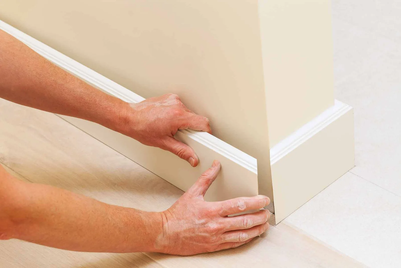 Hands fitting a length of baseboard trim into an inside corner above a plank floor