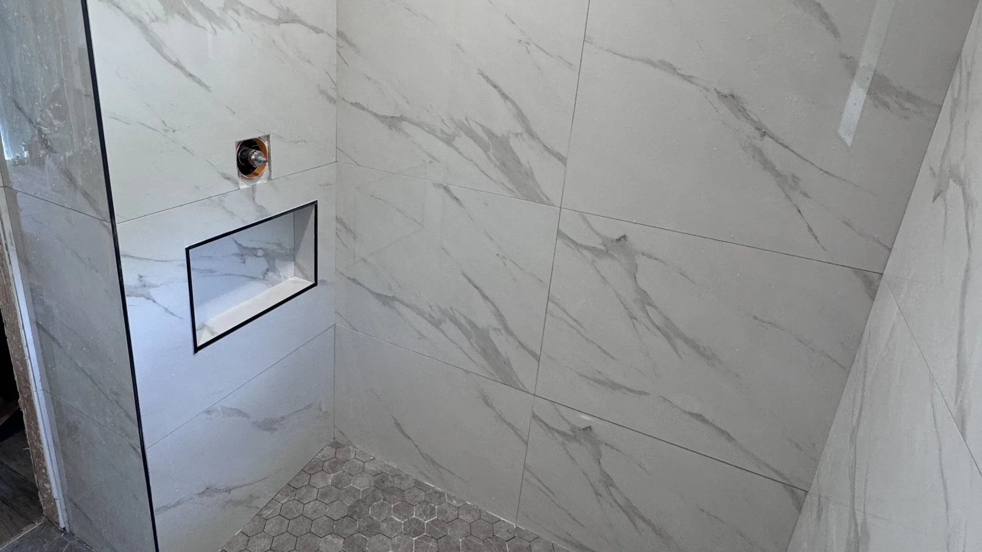 Walk-in shower in a St. Petersburg home tiled in large-format marble-look porcelain with a built-in niche and a grey hexagon mosaic floor