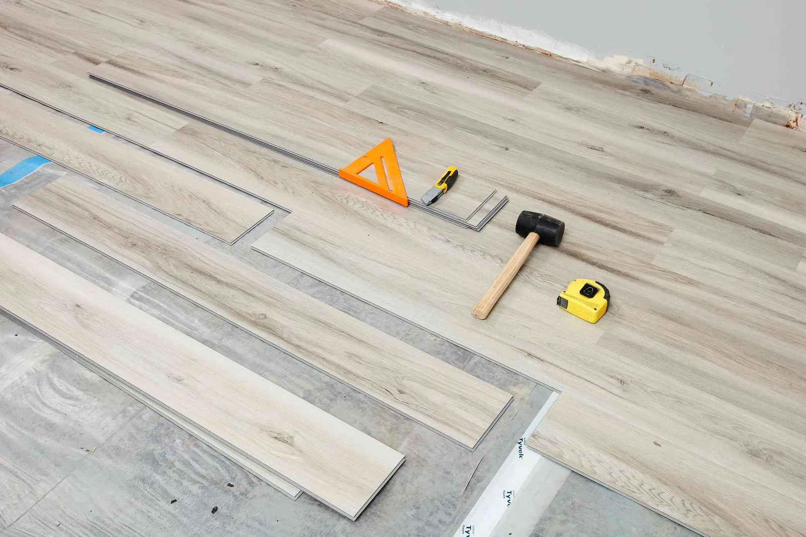 Vinyl plank flooring being laid with a tapping block, rubber mallet, speed square and utility knife on the floor