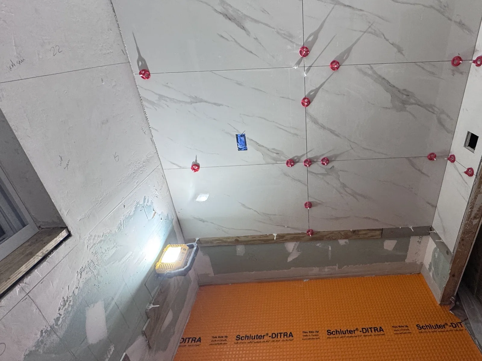 Large-format porcelain shower wall being set with red tile-leveling clips above an orange waterproofing membrane floor