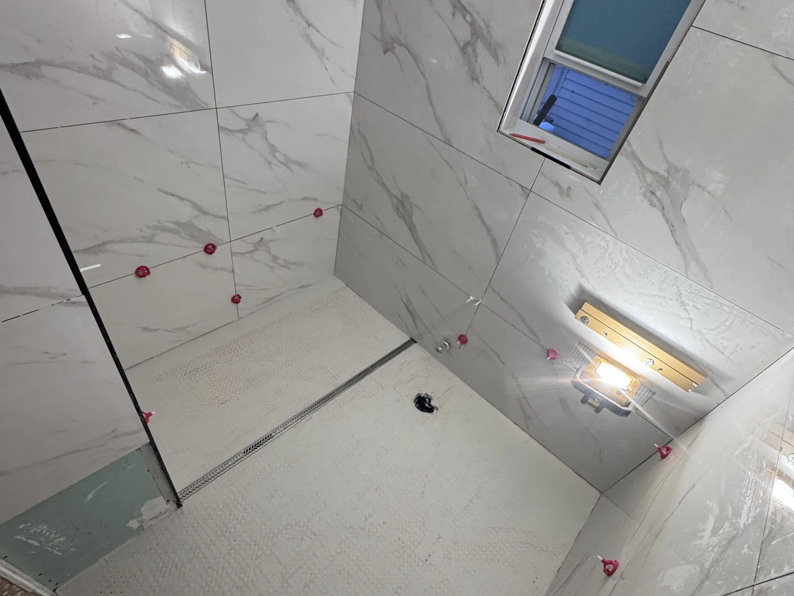 Large-format porcelain shower walls with leveling clips still in place, a white textured floor tile and a stainless linear drain