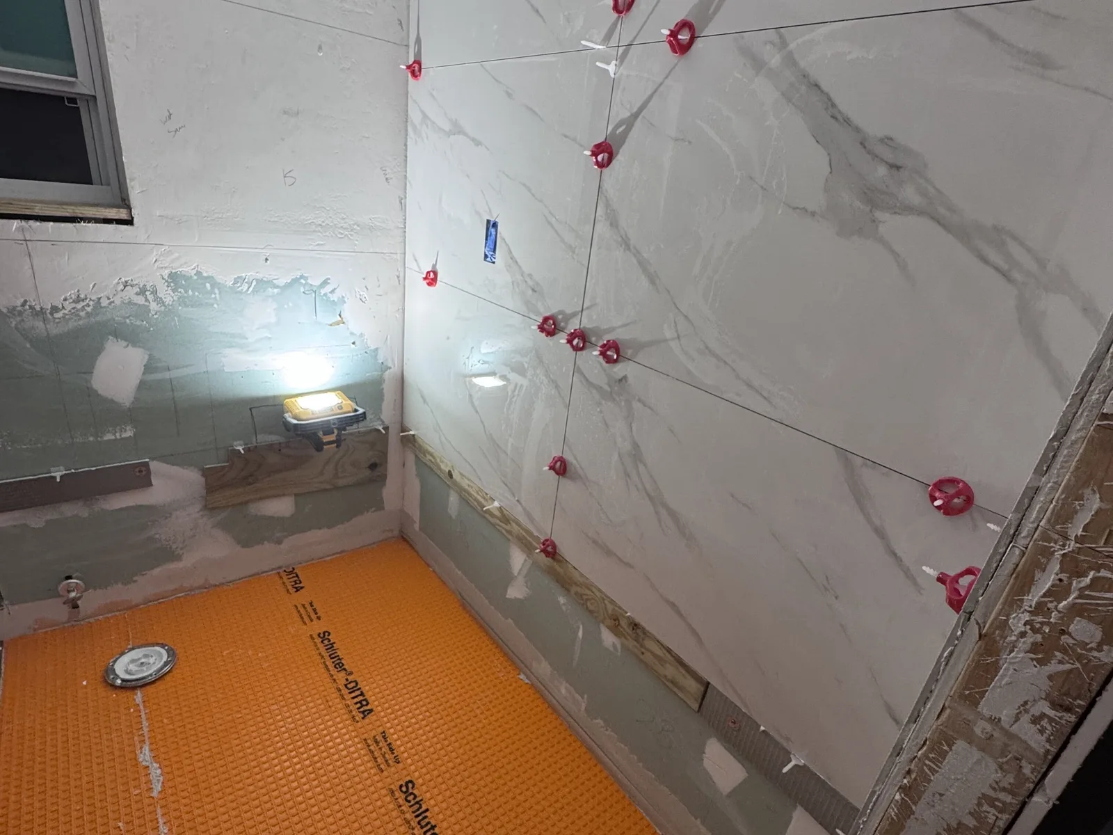 Tiled shower walls held by red leveling clips above a shower floor covered in an orange waterproofing membrane